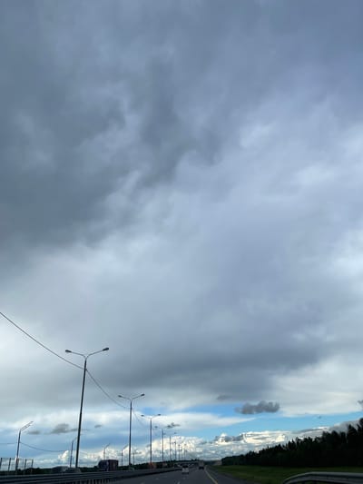 The image depicts an overcast sky dominating the left side, with a highway stretching into the distance on the right. A line of streetlights parallels the road. Trees line the far right edge. Vehicles are visible on the highway. The location is Бавыкино, Russia.