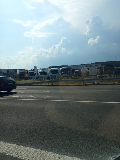 In Zvolen, Slovakia, a fleet of commercial trucks is parked in a lot, visible from a roadway. The trucks, primarily white with some orange and dark colored cabs, are arranged in a line. Several bear the "DAF" logo. One truck has a cylindrical tank, suggesting it's for liquid transport. A dark colored passenger car is parked to the left, facing away from the camera, with a license plate visible. The scene is set against a backdrop of a commercial building with signage, including "DAF STABO" and "ASTARO". A metal guardrail separates the roadway from the lot. The sky is partly cloudy, with dramatic white clouds against a bright blue sky. The ground is paved, with white lane markings visible on the roadway in the foreground. The time of day appears to be daytime, likely late morning or afternoon, given the lighting.