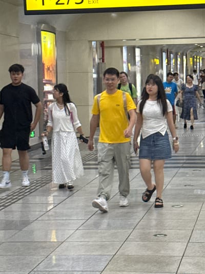 The image captures a group of people walking in a well-lit indoor environment, likely a transportation hub or a large public building in Longta, China, as indicated by a yellow sign at the top of the frame which reads "275 Exit". The flooring is made of large, light-colored rectangular tiles with dark grout lines, and there are circular metallic elements embedded in the floor at regular intervals.
In the foreground, a man wearing a black t-shirt and black shorts, white socks and white sneakers is walking from left to right. To his right, a woman in a white sleeveless top and a white skirt with blue polka dots walks in the same direction. She is holding a clear plastic bottle in her left hand and a dark object, possibly a phone, in her right hand. She is also wearing dark shoes.
Slightly ahead and to the right of the woman is a man wearing a bright yellow t-shirt, light gray pants, and white sneakers, also walking from left to right. Beside him, a woman in a white short-sleeved shirt and a blue denim skirt walks in the same direction. She is wearing dark sandals with straps. Her left wrist has a white watch, and her right wrist has a bracelet.
In the background, other individuals are visible, including a person in a blue t-shirt and another in a blue dress. The environment appears to be a corridor with vertical panels on the walls, some of which are illuminated with warm-toned advertisements or displays. The lighting is bright and even, suggesting an indoor public space. The overall scene depicts ordinary movement and activity within a city setting.