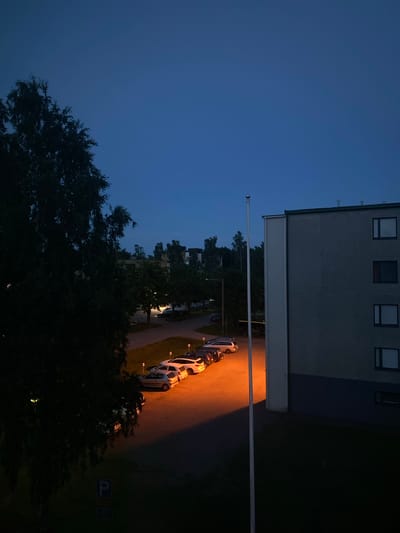 The image depicts a twilight scene in Kotka, Finland, likely in a residential area. On the right side of the frame is a multi-story building with a gray facade and several rectangular windows. A tall, slender pole extends vertically in front of the building. To the left, a large, dark tree dominates the foreground, obscuring much of the background. In the middle ground, a row of parked cars is illuminated by a bright, orange streetlamp. The street curves, and beyond it, more trees and buildings are visible, silhouetted against the darkening blue sky. The ground is paved in some areas and appears to be grass in others. There are no visible people or animals.