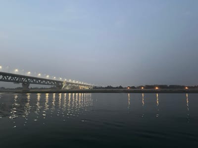 The image displays a wide-angle view of the Padma Bridge spanning the Padma River at twilight, near Naodoba, Bangladesh. The prominent feature is a long, multi-span truss bridge, dark grey in color, extending horizontally across the frame. Numerous streetlights along the bridge are illuminated, casting bright, elongated reflections onto the dark, calm water below. The bridge structure includes visible diagonal and horizontal members, supported by concrete piers. The far bank of the river, visible on the right, consists of a low-lying strip of land with sparse tree cover and a few scattered, distant artificial lights. The sky above is a uniform pale blue-grey, consistent with either dusk or dawn. No individuals, vehicles, or specific activities are discernable on the bridge or the water.