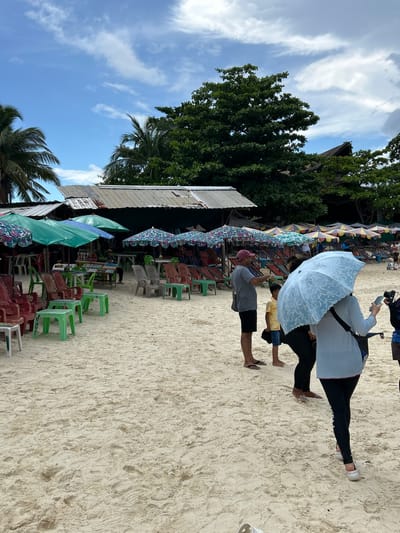 The image depicts a sandy beach scene with a row of open-air food stalls and seating areas under umbrellas. Several people are present on the beach. In the foreground, a person holds a light blue umbrella and a phone, with their back to the camera. To their left, a man in a grey t-shirt and shorts is looking towards the right, holding a phone. A young child in a yellow and blue patterned shirt stands between them. Further back, multiple rows of colorful beach umbrellas and chairs are set up. Behind the umbrellas and seating, there are structures that appear to be shops or restaurants with corrugated metal roofs. Lush green trees and palm trees are visible in the background, with a partly cloudy blue sky overhead. The scene is consistent with a beach resort or tourist area in Phru Nai, Thailand.