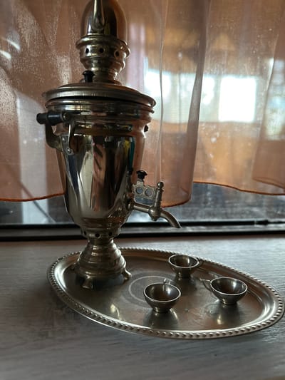 The scene is set indoors in Bol'shaya Mos', Russia. A polished, reflective metallic samovar, possibly brass or nickel-plated, occupies the central foreground. It features a decorative finial on its lid, a black-handled tap on its front right side, and a dark side handle on its left. The samovar's surface displays distorted reflections of the surrounding environment, including what appear to be humanoid forms or objects and various colors. The samovar rests on an oval, metallic tray with a distinct beaded edge. To the right of the samovar, on the same tray, are three small, dark metallic cups, each equipped with a handle. Behind the samovar, translucent, light-colored sheer curtains hang, partially obscuring a window that emits bright, diffused daylight. The curtains exhibit a textured appearance with illuminated particles. The objects are situated on a light-toned, textured surface, likely wood or laminate, suggesting a table or window sill. No individuals are visibly present or performing any actions.