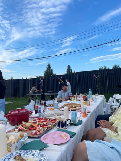 This outdoor scene, likely in Simagino, Russia, depicts a gathering at a long table laden with food and drinks under a blue sky with scattered clouds. Several people are present, some seated at the table and others standing. A man in a striped blue and white shirt is seated at the table, looking down, while another man in a light blue shirt stands in the background near a fence. To the left, a woman in a black top and light blue skirt stands facing away from the camera. A table tennis table is set up in the background, with one person playing.

The table is covered with a white tablecloth and is set with plates, cups, bottles of drinks, and various food items, including a large platter of cut vegetables and a red pot. The environment features a dark, vertical slatted fence in the background, with trees beyond it. The overall atmosphere appears to be a relaxed, outdoor social event.