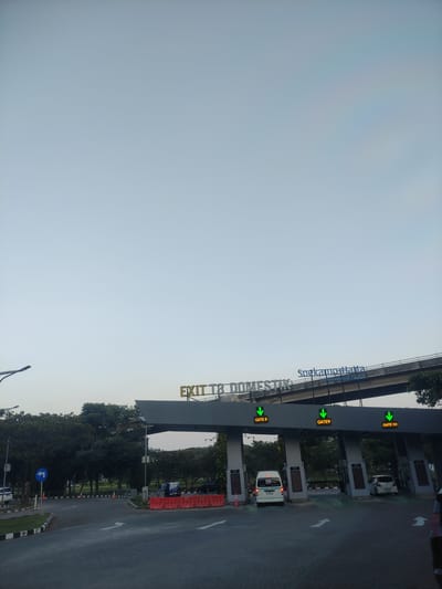 The image presents an outdoor, ground-level view of the domestic exit of Terminal 3 at Soekarno-Hatta International Airport in Rawa Burung, Indonesia.

In the foreground, an asphalt road with white directional arrows (indicating forward and left turns) leads towards a multi-lane gate structure. Black and white striped concrete curbs delineate the road edges. On the left side of the road, a blue circular sign with a white left-pointing arrow is visible, along with orange traffic cones and orange plastic water-filled barriers. A street lamp post is also present on the left.

Central to the image is a grey overhead gantry system spanning the width of the road. A prominent yellow-lettered sign on the gantry reads "EXIT T3 DOMESTIK". Above this, a blue sign displays "Soekarno-Hatta International Airport". Three distinct gate lanes are visible: "GATE 8", "GATE 9", and "GATE 10", each marked with an illuminated green downward arrow, signifying they are open. A white minivan is positioned directly under GATE 8, and a white compact car is visible under GATE 10. The front portion of a dark-colored vehicle is partially visible on the far right edge of the frame.

Above and behind the gate structure, a concrete overpass or bridge stretches horizontally across the scene. The background consists of dense green foliage from numerous trees lining the roadside. The upper two-thirds of the photograph is dominated by a clear, pale blue-grey sky, exhibiting a very faint, multi-colored spectral hue in the upper right quadrant.