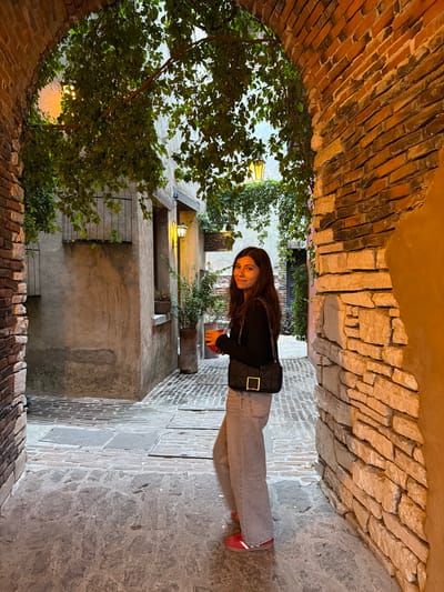 A young woman stands in a narrow alleyway in Domingo Arenas, Mexico. The alley is enclosed by brick walls and an arched brick ceiling. She wears a black long-sleeve shirt, light gray jeans, and red sneakers, and carries a black quilted bag. The alley floor is paved with stone. A tree with dense foliage is on the left side of the frame. In the background, a building with plants and a lit lantern is visible. The lighting is warm and subdued, suggesting the photo was taken in the evening.