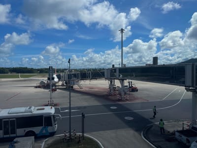 This is an image of an airport tarmac in Mai Khao, Thailand. In the foreground, a white bus with blue accents is parked. To the right, two individuals in high-visibility vests are walking on the tarmac. An airplane is connected to a jet bridge in the mid-ground. Various airport vehicles, including a red forklift, are also visible. The scene is set against a backdrop of a bright blue sky with fluffy white clouds, lush green trees, and distant buildings. The overall impression is a typical airport operational scene on a clear day.