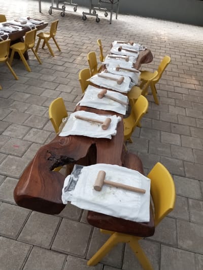 The scene depicts an outdoor or semi-outdoor paved area in Cibadak, Indonesia. The ground is covered with grey interlocking paver blocks. A prominent, elongated, irregularly shaped natural wood slab table, dark in tone, occupies the central midground. Around this table, several small, bright yellow chairs are positioned. On the table, seven individual stations are arranged, each consisting of a white rectangular item, possibly a package or placemat, upon which a small wooden mallet rests. Green particulate matter is visible on the white surfaces beneath several mallets. To the left, a portion of a smaller, rectangular wooden table with additional yellow chairs is visible. In the background, metallic structures on wheels are present against a light-colored wall or building facade. No individuals are present in the image.