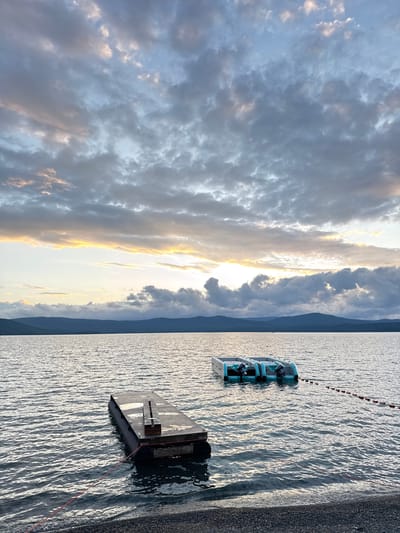 The image shows a scenic lake with a dramatic sky at sunset. The water is rippling and reflects the golden light from the sun breaking through the clouds. In the foreground, a dark, rectangular floating dock is partially submerged in the water, tethered by a red rope. Further out on the water, a light blue inflatable structure, possibly a water toy or a small boat, is also visible. The background features a distant mountain range silhouetted against the vibrant sky. The clouds are a mix of grays and whites, with streaks of gold and pink where the sun is shining through, indicating a beautiful sunset. The overall mood is serene and tranquil. The time of day is clearly sunset. No text is visible in the image.
