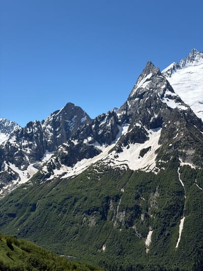 The image depicts a mountainous landscape under a clear blue sky. The foreground is dominated by a densely forested green slope that rises towards the middle ground. Above the tree line, the mountain slopes are covered with patches of snow and exposed dark rock. The peaks are jagged and sharp, with significant snow accumulation on the higher elevations. One prominent peak in the mid-right of the frame is particularly striking, with a steep, snow-covered face and a rocky summit. The overall scene conveys a sense of vastness and natural grandeur, characteristic of high-altitude mountainous regions. The location is in Верхне-Тебердинское сельское поселение, Russia, a region known for its mountainous terrain. No people or man-made structures are visible in the image.