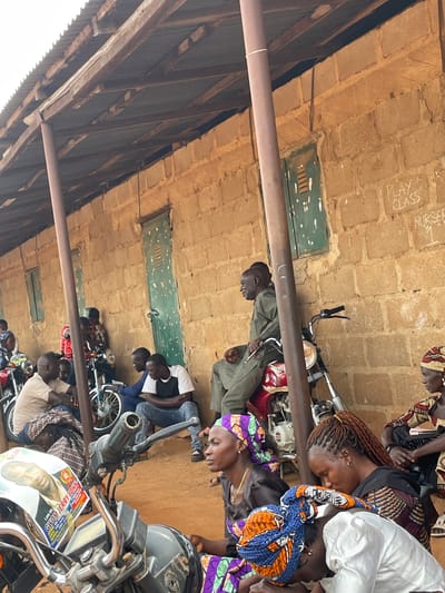 This image depicts a gathering of people outside a brick building with a corrugated metal roof supported by wooden beams and a metal pole. The wall of the building has "PLAY CLASS NURSERY 1" written on it in chalk. Several motorcycles are parked in front of the building.

In the foreground, a motorcycle's handlebars and part of its body are visible. A flyer featuring the face of a man named "GWOM JERRY JOHN" is attached to the motorcycle.

The people in the image are dressed in casual clothing and headscarves. Some are seated on the ground, while others are standing or sitting on motorcycles. Their interactions appear to be casual, with some conversing and others looking in different directions. The overall atmosphere seems relaxed, with the setting suggesting a community gathering or a temporary stop.

The scene is likely located in Kassa, Nigeria, as indicated by the request. The environment appears to be outdoors, with dusty ground and a simple, functional building. The lighting suggests it is daytime.
