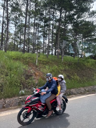 The image shows two people riding a red motorcycle on a paved road. The rider in the front is wearing a blue jacket, brown pants, and a dark helmet with a white face mask. The passenger behind is wearing a grey jacket, patterned pants, and a white helmet with a white face mask. The motorcycle is a Yamaha Exciter, identifiable by its red and black color scheme and decals. The background features a grassy slope with scattered trees, predominantly pine trees, suggesting a rural or semi-rural environment, possibly on the outskirts of a city like Thôn 6, Vietnam, where such landscapes are common. The road has a white line marking its edge. The lighting suggests it is daytime, with overcast skies. The overall scene depicts casual transportation through a scenic, green landscape.