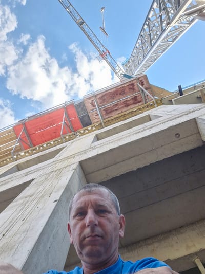 The image depicts a low-angle perspective of a construction site with two cranes in the background against a bright blue sky with scattered clouds. In the foreground, a man in a blue shirt is looking towards the camera. He has short, graying hair and is facing slightly upwards. Part of a concrete structure, likely a building under construction, is visible to the right of the man and extends upwards. Scaffolding with orange and brown planks is attached to the side of the concrete structure. The image appears to have been taken during the daytime, indicated by the bright sky and sunlight. The overall scene suggests an active construction environment. No visible text is present in the image.