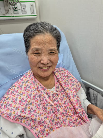 An elderly East Asian woman with short, graying hair and freckled skin smiles at the camera. She is sitting in a hospital bed, leaning against a light blue pillow that has Korean text printed on it, which appears to say "SUWON HYO HOSPITAL" and "수원효병원". She is wearing a light-colored hospital gown and a pink bib with a floral pattern of small pink, blue, and yellow flowers. The background shows hospital equipment, including medical gas outlets, and a wall. The image appears to have been taken in Suwon-si, South Korea, based on the text on the pillow.