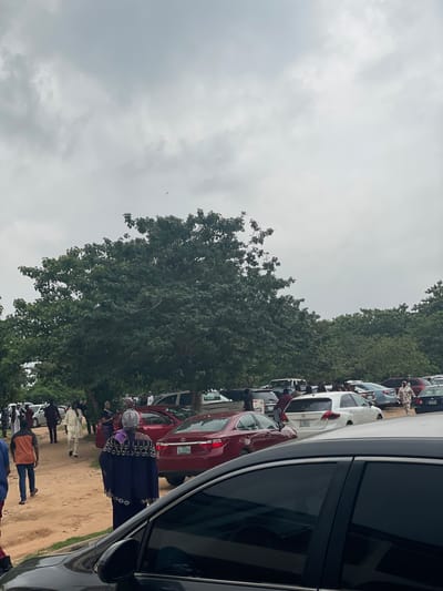 An outdoor scene in New Nyanya, Nigeria shows a gathering of people and parked vehicles under a cloudy sky. The ground is a mixture of dirt and sand. Several trees with dense green foliage are visible, some of which are large and mature. A variety of cars, including sedans and SUVs, are parked in an open area.
People are seen walking around and interacting. Some individuals are dressed in traditional Nigerian attire, including flowing robes and head coverings. One person is wearing a patterned tunic and trousers, and another is in a dark blue garment with intricate embellishments. A man in a light-colored outfit is walking away from the camera, while others are seen in small groups. The overall atmosphere suggests a community event or a gathering.