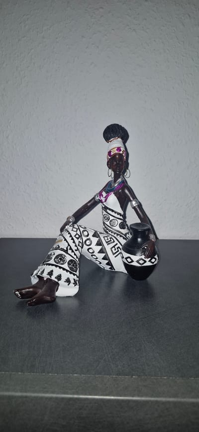 A dark-skinned figurine of an African woman sits on a dark surface against a textured, light-colored wall. The woman is depicted wearing a white and black patterned garment, a white headdress with colorful accents, and jewelry. She is holding a black pot with a patterned band. The figurine is in a seated position with her legs extended. The background appears to be a wall in a room. The location is not specified as Baden-Baden, Germany.