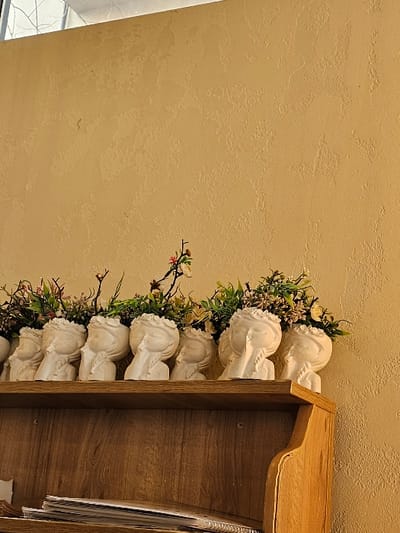 The image displays an interior scene in Fundeni - Dobroești, Romania. A row of at least eight white, ceramic head-shaped planters, depicting female faces with closed eyes and hands near the chin, rests on the top surface of a light brown wooden shelving unit. Each planter contains a mix of artificial green foliage and small, light-colored flowers. On a lower shelf of this unit, multiple stacks of paper or notebooks are visible. The background consists of a uniformly textured, light yellow-beige wall. A white ceiling or structural element is partially visible in the upper left corner of the frame. No individuals are present.