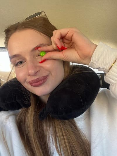 A female subject is depicted in a close-up portrait, seated within what appears to be the interior of a vehicle. She has long, light brown hair and is wearing a light-colored, long-sleeved top and a black U-shaped travel neck pillow. Sunglasses are resting on top of her head. Her right hand, with fingers painted with red nail polish, holds a single spherical green candy between her thumb and index finger. This candy is positioned in front of her partially closed left eye, creating a playful illusion. Her right eye is open, and her lips are curved into a slight smile, accentuated with reddish-brown lipstick. The background is a plain, light-colored interior wall or ceiling, characteristic of a vehicle's cabin. The specific city of Jasenovo Polje, Montenegro, or any other precise geographical location, is not discernible from the visual information provided.