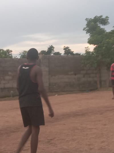 A person in a black tank top and shorts is walking on a dirt path in Achara Ndiagu, Nigeria. A cinder block wall runs parallel to the path, with trees and sky visible in the background. Another person can be seen in the background with a striped shirt.