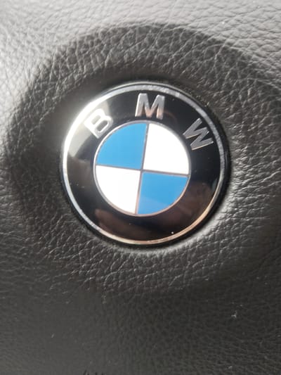 The media file shows a close-up view of a BMW logo on a black, textured surface, likely a car's steering wheel or dashboard. The BMW emblem is circular with a metallic silver rim. Inside the rim, the logo is divided into four quadrants. Two opposing quadrants are a bright blue, and the other two are white. The letters "B", "M", and "W" are prominently displayed in metallic silver across the top portion of the emblem. The surrounding surface is black and exhibits a distinct leather grain pattern. The lighting suggests it is daytime, and there are no immediate indications of the specific location within Krchleby, Czechia, beyond the general context. The image is focused on the logo, with the background slightly out of focus, emphasizing the brand. No people are visible in the frame.