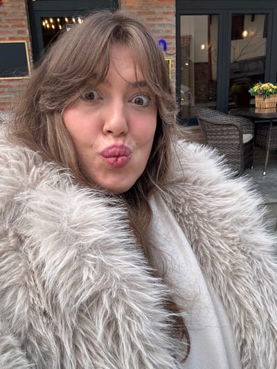 A light brown-haired female subject is depicted in a close-up, exhibiting a wide-eyed expression with puckered lips. She is dressed in a light grey shaggy faux fur coat, layered over a cream-colored garment. The background indicates an outdoor cafe environment located in Sighnaghi, Georgia. Visible elements include a dark wicker chair with a light cushion and a round, dark-colored table. A wicker basket containing yellow and purple artificial flowers rests on the table. Behind this setup, a brick building facade with a dark-framed glass door reflecting interior lights is present. The overall lighting is subdued.