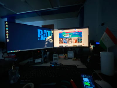 The image depicts a dimly lit desk setup with two computer monitors. The left monitor displays a dark background with the word "BATMAN" in large, glowing blue letters. The right monitor shows a webpage with various product advertisements and promotional banners, including "LOVI PODARKI OT SBERA" which translates to "Catch Gifts from Sber". On the desk, there are several objects including a keyboard, a mouse, a mobile phone displaying what appears to be a stock market app, a bottle of what looks like perfume or cologne, a pair of glasses, and a small flag of Tajikistan. The environment appears to be an indoor workspace, possibly an office or a home office, given the presence of the computers and desk accessories. There is no indication of the city of Yukhta, Russia, in the image.