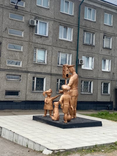 A golden bronze sculpture depicts a man in a postal uniform, possibly a postman, standing next to a traffic light signal that appears to be a part of the sculpture. He is interacting with two children and a dog, all also rendered in golden bronze. The scene is set against a backdrop of a gray, multi-story apartment building with numerous windows and air conditioning units. The building's facade is textured and appears to be made of concrete panels. The sculpture is situated on a raised concrete platform paved with square tiles, surrounded by a low, damaged concrete curb and overgrown grass. The overall setting suggests an urban environment in Usolye-Sibirskoye, Russia. The children are positioned at the man's feet, one reaching out towards him, while the dog sits beside them. The man appears to be addressing the children, and the traffic light is positioned behind him. The image is taken from a low angle, emphasizing the sculpture and the building.