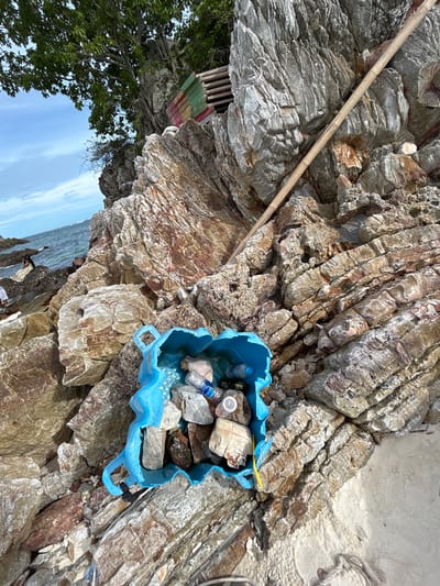 This image depicts a rocky shoreline with a blue plastic container filled with trash, including plastic bottles and other debris. The container is situated on a beach with large rock formations. In the background, the ocean is visible, and several people are present on the rocks. A large tree with green foliage grows from the rocky cliff, and a bamboo pole leans against the rocks. Colorful wooden planks are stacked on the cliff face above the tree. The scene is likely located in Tambon Phru Nai, Thailand, given the characteristic rocky coastline and tropical vegetation.