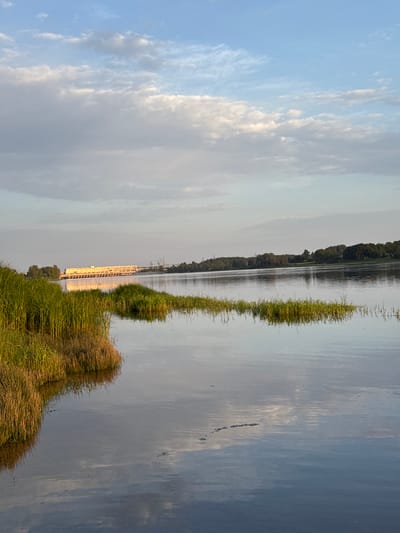 A wide, calm body of water, likely a river, dominates the foreground and mid-ground of the image. The water reflects the soft hues of the sky and the surrounding landscape. On the left bank, dense green and golden reeds grow from a small island or peninsula, casting a shadow onto the water. In the distance, across the river, a long, low structure with numerous windows and a yellow-tinted facade is visible, possibly a dam or a large industrial building, illuminated by the setting sun. Beyond this structure, a line of trees forms the horizon, and above them, a pale blue sky is adorned with scattered, wispy clouds, some tinged with the warm light of sunset. The scene appears serene and natural, with a hint of industrial presence in the background. This location is identified as Rumbenieši, Latvia, suggesting the river could be the Daugava.