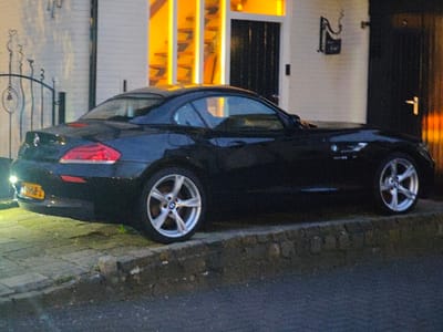 A black BMW Z4 Coupe (E86 model) with silver multi-spoke alloy wheels is stationary, viewed from its rear three-quarter aspect, under ambient low light conditions consistent with evening or night. The vehicle displays a yellow Dutch license plate, '7-P6B-Z', affixed to its rear. It is positioned on a paved surface that is elevated above a darker asphalt road, separated by a low, dark-colored stone wall. The car is parked adjacent to a building featuring a light-colored facade. Visible architectural elements include a dark main entrance door to the left, and a secondary dark door further right. Orange light emanates from within the building or from decorative panels near the main entrance. A dark, ornate wrought iron gate with a bell is visible to the left of the vehicle's rear, and a wall-mounted decorative fixture is present on the facade between the two doors. No individuals, actions, or interactions are depicted. The scene is located in Wierden, Netherlands.