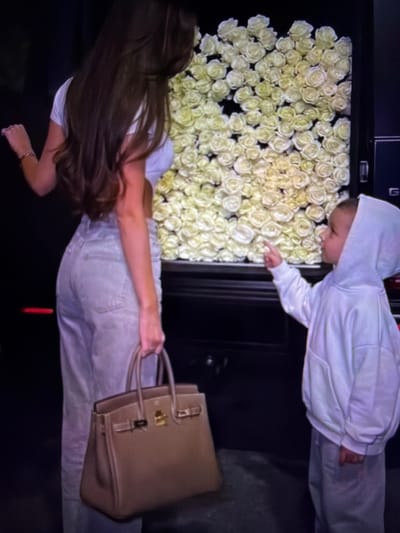 In this media file, a young boy in a white hoodie and light-colored pants is looking at a wall covered in cream-colored roses. He is pointing at the roses. Next to him, a woman with long brown hair is wearing a white t-shirt and light-colored pants. She is holding a tan-colored handbag. The scene appears to be indoors, with a dark background. The image has a vertical orientation, with the subjects appearing to be viewed from above and from the side. The lighting is dim, suggesting it might be evening or an interior with artificial lighting. There is no visible text in the image. The overall mood is calm and observational.