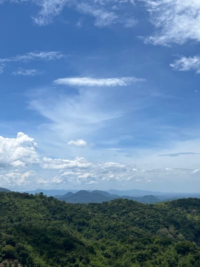 The image depicts a panoramic view of lush green mountains under a bright blue sky with scattered white clouds. The foreground is dominated by a dense forest covering rolling hills. In the background, several layers of mountains recede into the distance, becoming progressively bluer and hazier, suggesting a vast landscape. The sky is a vibrant blue with wispy white clouds, and larger, puffier clouds are visible on the left side, indicating fair weather. The scene appears to be a natural landscape, possibly a viewpoint overlooking a mountainous region. This specific landscape could be found in the rural or mountainous outskirts of Son Dien, Vietnam, known for its scenic natural beauty and hilly terrain.