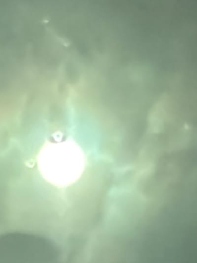 The image displays a bright, circular light source, strongly resembling the sun, seen through a heavily diffused and overcast sky. The atmosphere is characterized by a muted green-grey color palette, suggesting dense cloud cover or significant atmospheric haze. Indistinct, soft cloud formations are visible, scattering the light and creating a soft, ethereal quality.
Surrounding the central bright light, several smaller, irregular bright anomalies or lens flares are visible, indicating reflections within the camera's optics or atmospheric phenomena. The image is somewhat blurry, lacking sharp detail, which contributes to the abstract and atmospheric nature of the scene.
There are no discernible people, specific man-made objects, or visible text. The scene depicts an outdoor sky under overcast conditions. Given the context of Lørenskog, Norway, this image captures the daytime sky above that location. No specific activity or event is taking place beyond the observation of the sky and sun. The time of day is daytime, though the diffused light makes it difficult to pinpoint morning, midday, or afternoon. No specific emotions are conveyed.