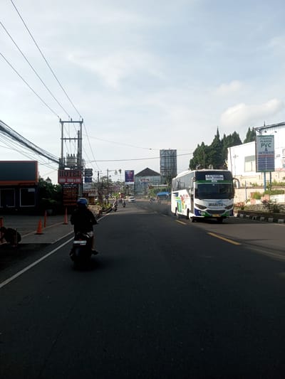 The image depicts a street scene in what appears to be a city with a mix of commercial and residential areas. On the left, there's a restaurant or hotel with signs reading "LUXURY GARDEN VIEW" and "SHABU-SHABU & GRILL", advertising amenities like a fitness gym and swimming pool. Orange cones line the curb, and a person is working near the edge of the road. A motorcyclist rides down the street, and a bus is traveling in the opposite direction. Several billboards and signs are visible, including an advertisement for "Gemilang PLAFON PVC & INTERIOR". Power lines crisscross above the street. Other motorbikes are on the street in the distance. The sky is partly cloudy. There is no indication that this is a city of Phou Khoun, Laos.
