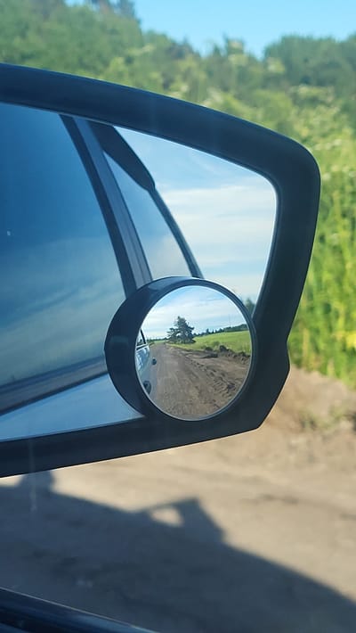 The image is a close-up shot of a car's side mirror, reflecting a dirt road with a single tree in the distance. The background shows a green field under a blue sky with some clouds. The car itself is not fully visible, but a shadow of it is cast on the ground. The scene is set in Ural'skoe, Russia.
