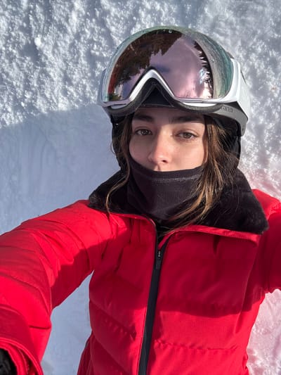 A person lies on snow wearing a red ski jacket and a black neck gaiter. Ski goggles with a reflective lens rest on their head, reflecting trees and sky. The person looks at the camera with brown eyes. The snow is white with slight shadows. The jacket is zipped up and has a black zipper and black fur lining. The person's brown hair is visible.