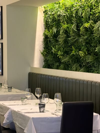 The image depicts an indoor dining setting in Otranto, Italy, featuring a table laid for a meal. White tablecloths cover the tables, which are set with glassware, cutlery, and decorative elements. A prominent feature of the background is a lush, vertical garden wall, creating a natural and vibrant atmosphere. The seating area includes a dark upholstered bench and chairs. The lighting appears to be artificial, casting a warm glow on the scene. The overall impression is one of a sophisticated and inviting restaurant interior.
