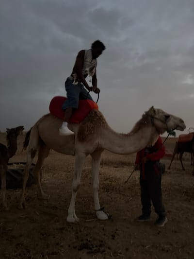 A person rides a camel in a desert landscape under a cloudy sky. The rider, wearing a patterned vest and blue jeans, is positioned on a red saddle. Another person, dressed in a red top and dark pants, stands beside the camel, holding a lead rope. In the background, another camel is visible, also with a saddle. The ground is sandy and dry, with some sparse vegetation. The overall lighting suggests it is dusk or dawn. The location is identified as Douar Ait Daoud, Morocco.