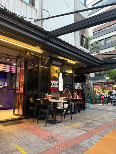 The image shows a street-level view of a burger restaurant with outdoor seating in İzmir, Turkey. The restaurant has a modern design with a black awning, glass doors, and yellow neon lighting. Inside, a person in a blue cap is visible behind the counter. Outside, there are several tables and chairs where customers are seated. A neon sign on the wall reads "BURGER IS THE ANSWER". In the background, there are buildings, trees, and a street with a yellow taxi and a scooter. The pavement is paved with grey and red tiles, and yellow lines are painted on the ground, possibly indicating a walkway or social distancing markers. The overall impression is a bustling urban scene during daytime.