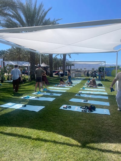 The scene depicts an outdoor fitness event in Thermal, United States, under a white canopy structure. Attendees are spaced on light blue yoga mats arranged in rows on a green lawn. Some people are engaged in physical activity, while others observe or walk around. Exercise equipment like kettlebells and small weights are placed on the mats. In the background, there are palm trees, other canopy structures, and a colorful mural. The sky is clear and blue.