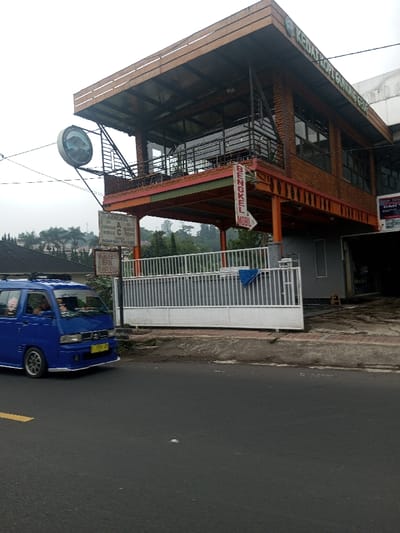A two-story building with a coffee shop named "KEDAT KOPT GUNUNG GE808" visible on the upper level. A sign indicates "BENGKEL MOBIL" (mobile workshop). Additional signage mentions "BENGKEL" with details on air conditioning services, and "NITROGEN BAN TUBELESS." A blue van drives along the road in front of the building, alongside a white fence.