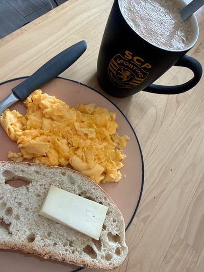 A breakfast scene is depicted on a wooden table, likely in Almada, Portugal, given the mug's logo. A black mug with the "SCP Sporting Portugal" crest contains a frothy beverage, possibly coffee or hot chocolate. To the left of the mug is a light pink plate holding a generous portion of fluffy, yellow scrambled eggs. Beneath the eggs and slightly to the left is a slice of rustic bread with a light brown crust and a porous interior. A small, rectangular piece of white cheese sits atop the bread. A dark-handled knife with a matte finish rests on the plate, angled towards the eggs. The wooden table surface displays a natural grain pattern.