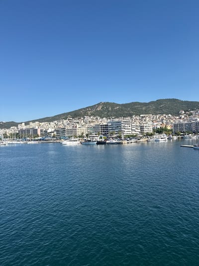 This image shows a view of the harbor and waterfront of Kavala, Greece. In the foreground, the blue water of the harbor is calm with small ripples. Moored boats of various sizes, including yachts and sailboats, are visible along the waterfront. Behind the boats, a strip of land features buildings, ranging from modern structures with white facades to older, more traditional architecture. These buildings climb up the side of a hill that forms the backdrop of the city. The hill is covered in greenery and also dotted with buildings. The sky above is a clear, bright blue, indicating a sunny day. The scene is one of a bustling port city situated against a scenic natural landscape.