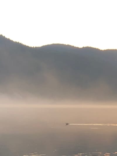 The image shows a serene, hazy landscape with a body of water in the foreground and a misty mountain range in the background. The sky is a bright, washed-out white, indicating either dawn or dusk. The water is calm and reflects the soft light, with a faint glimmer on its surface. In the middle distance, a small boat with two figures is visible, leaving a thin white wake as it moves across the water. The mountain range is shrouded in mist, obscuring its details and creating a sense of depth and atmosphere. The overall impression is one of tranquility and natural beauty. The location is identified as Слизнево, Russia, suggesting a rural or natural setting.