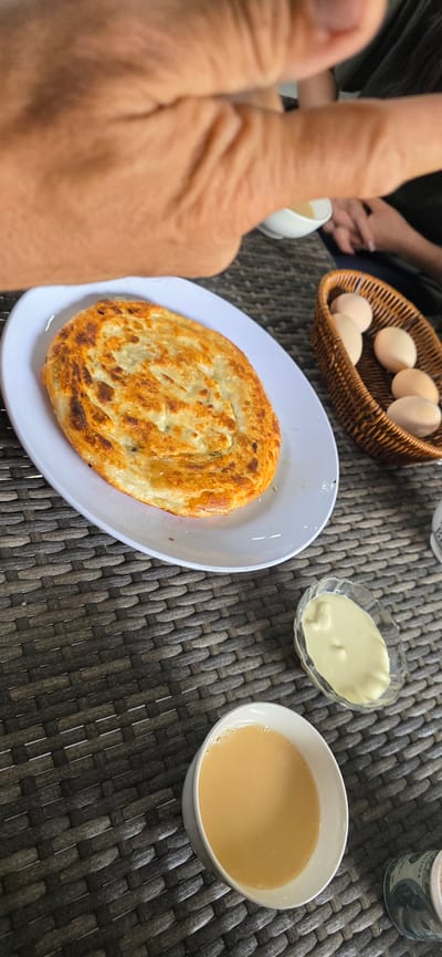 A close-up, angled shot shows a table setting with food and beverages. In the foreground, a white plate holds a golden-brown, layered flatbread. To the right of the flatbread, a small glass bowl contains a pale yellow, creamy substance. Further to the right, a woven basket holds several light brown eggs. A white ceramic mug filled with a light brown liquid, possibly tea, is positioned in the lower right quadrant of the image. The table surface is a dark woven material. In the upper portion of the image, a person's hand, with visible wrinkles, is partially visible, suggesting someone is reaching for or interacting with the food. In the background, out of focus, another person's hands and arms can be seen. The environment appears to be outdoors, possibly a cafe or restaurant in Sasyk-Bulak, Kyrgyzstan.