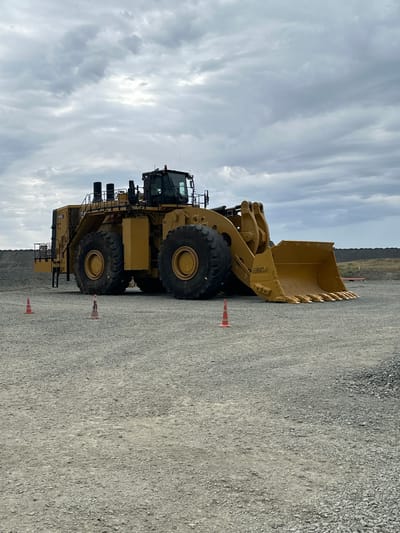 A massive, yellow Caterpillar 995 wheeled loader sits idle on a gravel surface under an overcast sky. The loader is positioned in the foreground, with its large bucket facing the viewer's right. Its immense tires dwarf the two orange traffic cones placed in front of it. In the background, a large pile of earth or rubble is visible, suggesting a mining or construction site. The sky is filled with dense, grey clouds, typical of the weather in Bozshakol, Kazakhstan, known for its mining operations. There are no people or active operations visible in the image.