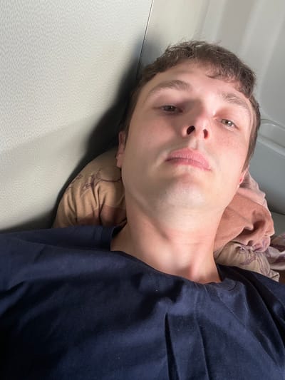 A young man with light brown hair, freckled skin, and brown eyes is lying down, looking upwards at the camera. He is wearing a dark navy blue t-shirt. His head is resting on a patterned pillow, which is placed against a textured, light beige surface that appears to be a wall or upholstery. The background is out of focus, but suggests an indoor environment, possibly a vehicle or a room. The lighting is soft and diffuse. No specific location in Sady, Poland can be determined from the image alone.