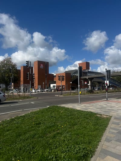 The image captures an exterior view of Stevenage Railway Station in Stevenage, United Kingdom, under a clear blue sky with scattered white cumulus clouds. The prominent structure is a multi-story building constructed from red brick, featuring the red National Rail double arrow logo and "Stevenage" text on its central section.
In the mid-ground, an elevated grey pedestrian footbridge with stairs extends from the station building, spanning over an asphalt car park area. Several vehicles, including white and dark-colored cars, are parked within this area. A transparent, arched bus shelter is visible on the left side of the car park, closer to the station entrance. A black metal fence runs along the edge of the car park, separating it from the main road.
In the foreground, a two-lane asphalt road with white lane markings occupies the left side, with a silver vehicle partially visible on the far left, moving away from the viewer. To the right of the road, a paved pedestrian pathway constructed of rectangular grey and reddish-grey blocks borders a patch of green grass containing some dandelions. Multiple dark grey poles support traffic light arrays at what appears to be a road intersection or crossing. One traffic light on the right displays a red signal. No individuals are prominently visible engaged in specific actions or interactions within the scene.