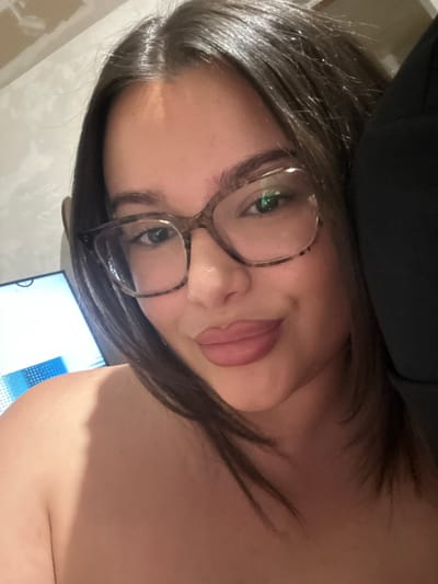 A close-up, front-facing selfie shows a young woman with dark brown hair and glasses. She is wearing light pink lipstick and has a fair complexion. The background appears to be an indoor setting with a light-colored wall and a dimly lit area to her right. A monitor screen is partially visible to her left, displaying a blue and white pattern. The woman is looking directly at the camera. There is no indication of the specific location within Vaglen, Bulgaria.