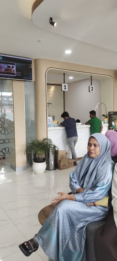 The image depicts the interior of a bank or financial institution. In the foreground, a woman wearing a blue hijab and a matching patterned dress is seated on a chair, looking to her left. She is wearing black sandals. Behind her, two men are standing at a counter. One man, wearing a striped blue and grey shirt and grey pants, is facing away from the camera, leaning on the counter. The other man, wearing a green t-shirt and dark pants, is also facing away and appears to be interacting with someone at the counter. In the background, a counter area is visible with signage indicating service numbers "1" and "2". A decorative patterned wall is behind the counter. To the left of the counter, a large screen displays banking information, including a queue number "B055". A potted plant is situated on the floor next to the counter. Cardboard boxes are stacked on the floor to the right of the plant. The floor is tiled with a marble pattern. The overall lighting appears to be from overhead fixtures and the screen. The location is identified as Keudealue Ie Puteh, Indonesia.